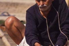 A shaven headed man sits crosslegged at the Ganges riverside, making offerings