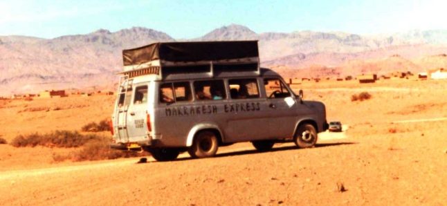 A minibus with large roof rack for camping gear speeds through the sandy terrain to the south of the Atlas mountains