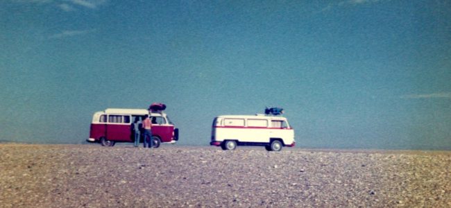 Two volkswagen campervans are apparently perched exactly on the horizon where a dry stony desert plain meets the unblemished skies
